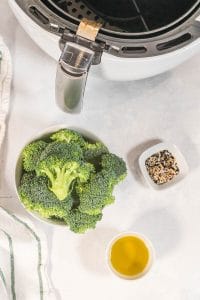 Vertical photo of ingredients for Everything But The Bagel Broccoli. Broccoli florets, olive oil and Everything But The Bagel mixture. In the background there is the basket of a white air fryer. The basket has a silver handle.