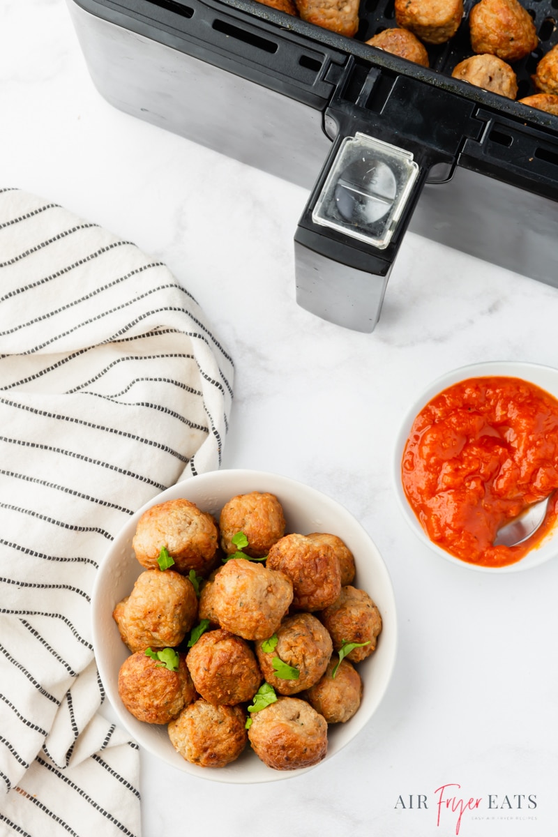 a white bowl filled with cooked meatballs, viewed from above next to an air fryer and a bowl of marinara sauce