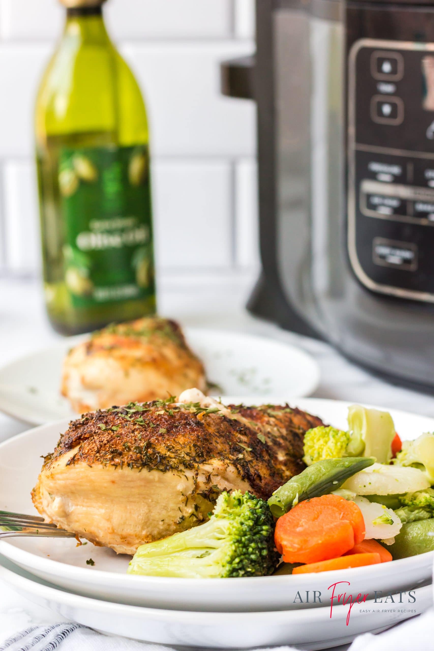 a dinner plate of air fried chicken breast and frozen vegetables in front of a Ninja Foodi and a bottle of olive oil