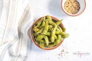 Horizontal photo of a dish containing cooked air fryer edamame. There is a smaller bowl containing salt & pepper. At the bottom of the image is the company logo