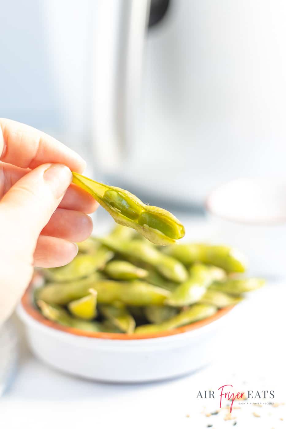 A vertical photo of a white dish containing air fried edamame. In the background is a white air fryer, and just to the left is a hand holding a partially split edamame pod. To the bottom right of the image is the company logo