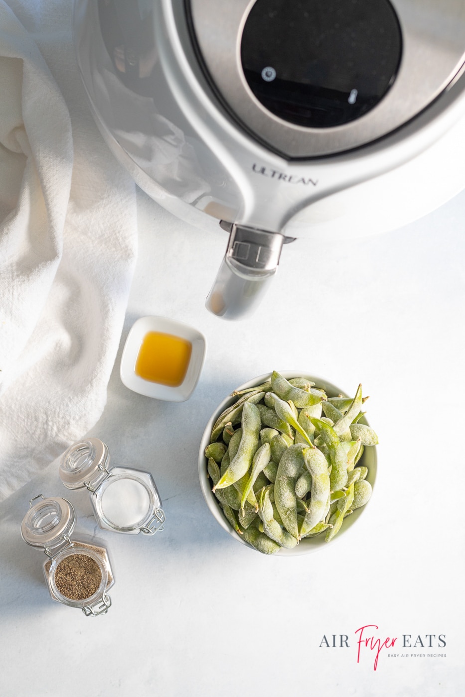 Vertical photo of ingredients required for Air Fryer Edamame. At the top of the photo is a white air fryer. There is a bowl containing frozen edamame, salt and pepper in pots and a tub containing sunflower oil. To the bottom right of the photo is the company logo