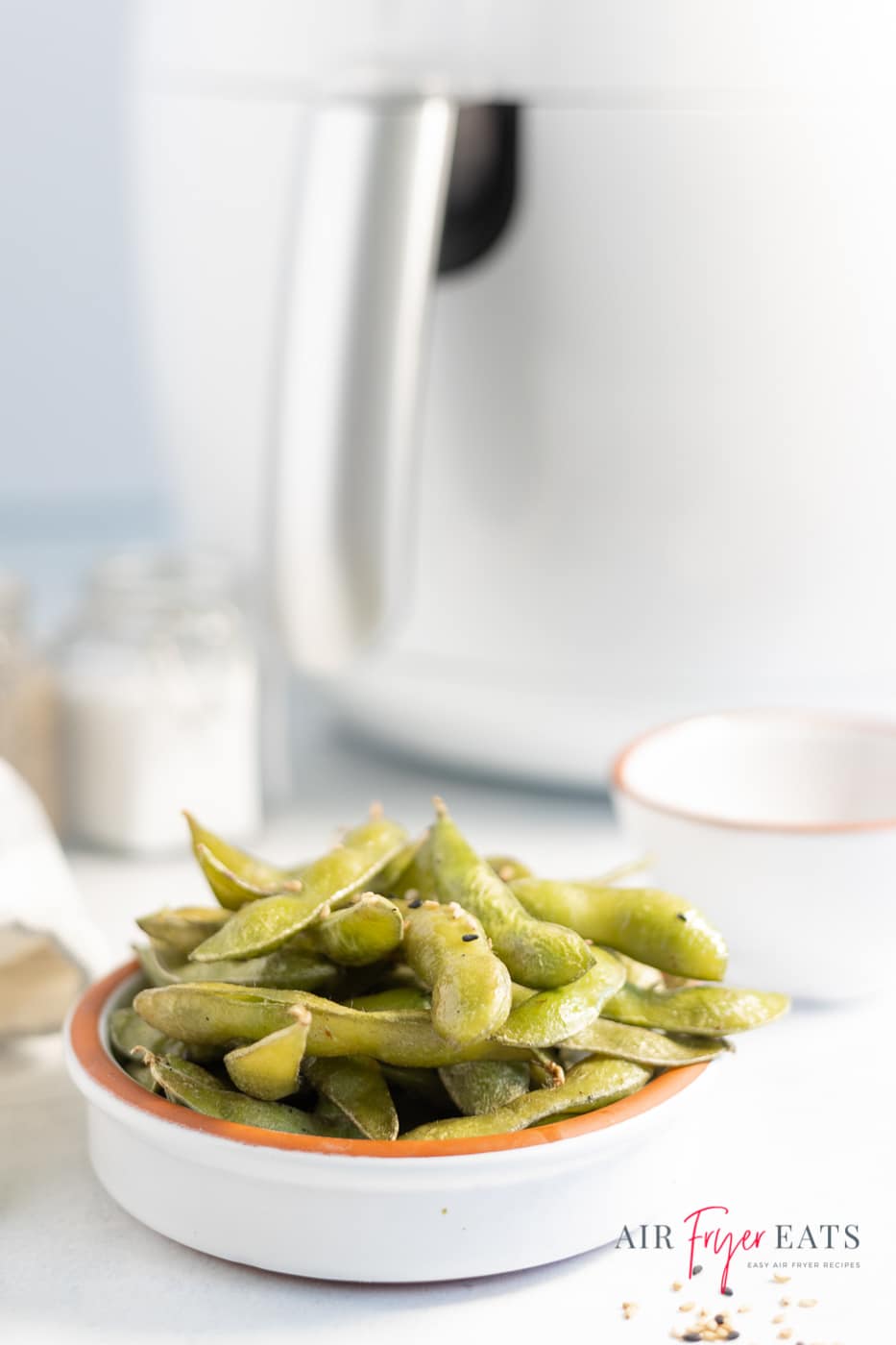 Vertical photo of a white dish containing air fryer edamame. In the background is a white air fryer. At the bottom of the photo is the company logo