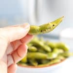 Close up vertical photo of a hand holding an air fried edamame pod. In the background there is a white bowl containing edamame pods. There is also a white air fryer. The company logo is at the bottom of the image