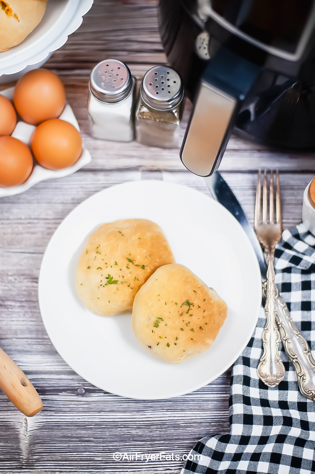Overhead shot of two egg and bacon stuffed biscuits on a white plate. There is a black air fryer in the background with a silver handle, a packet of eggs and a knife & fork to the right of the plate