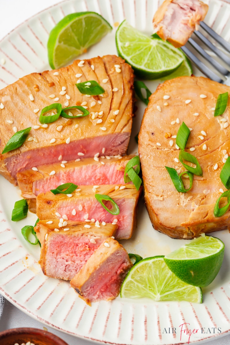 a ridged white plate holding two air fryer tuna steaks, garnished with lime wedges, sliced green onion, and sesame seeds