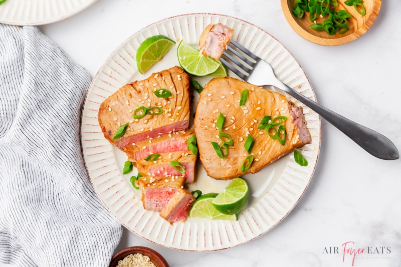 horizontal image showing a dinner plate filled with air fried tuna steaks. One has been sliced. There are lime wedges on the plate and a silver fork is holding a piece of fish. 