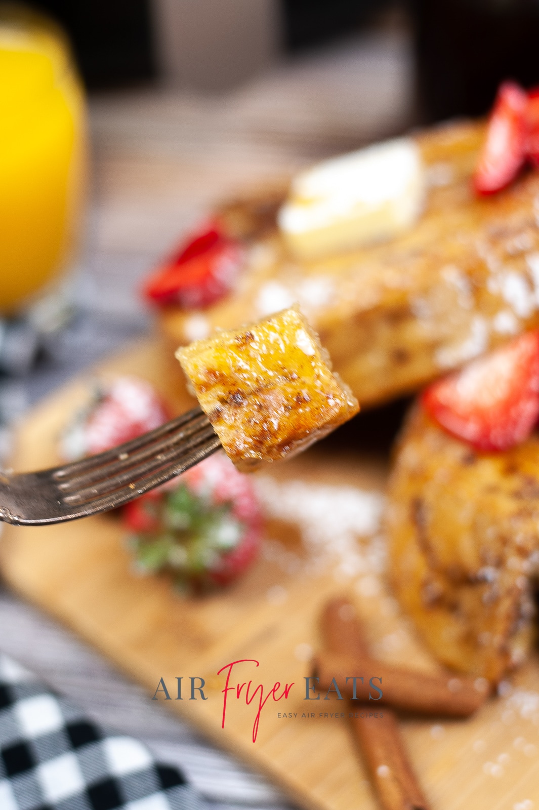 A fork with a bite of air fryer french toast being lifted up. In the background is a board with french toast with strawberries