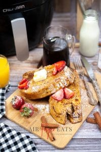 A vertical photo of air fried french toast on a wooden board with strawberries and a knife & fork. In the background there is a bottle of milk and a black air fryer with silver handle
