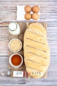 Vertical photo of ingredients required to make air fryer french toast. French bread loaf, milk, eggs and spices.