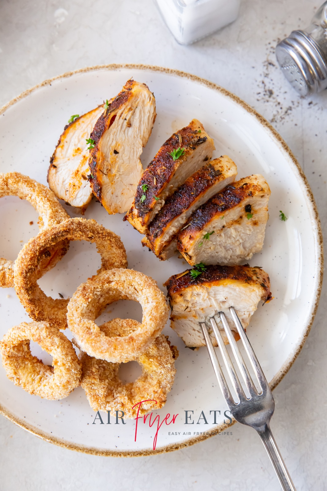 chicken breast sliced on a white plate with green herbs on top with a fork stuck in one piece and onion rings to the side