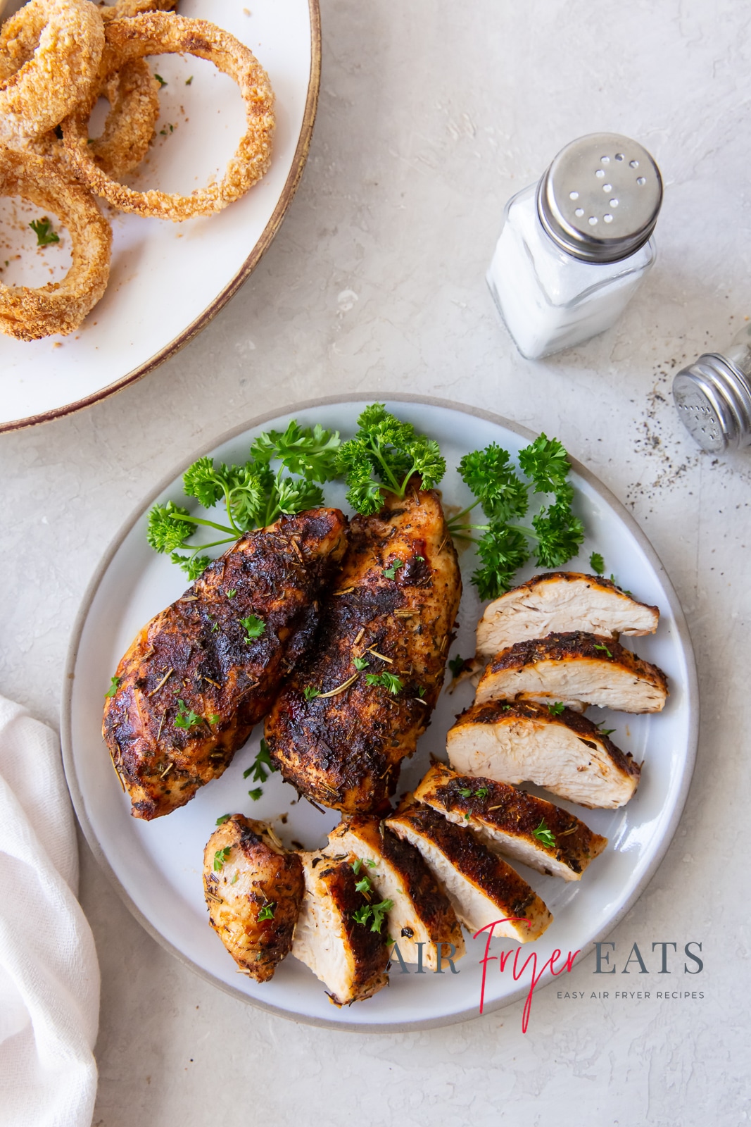 chicken breast sliced on a white plate with green herbs on top and to the side. salt and papper holders to the top right and onion rings to the top left