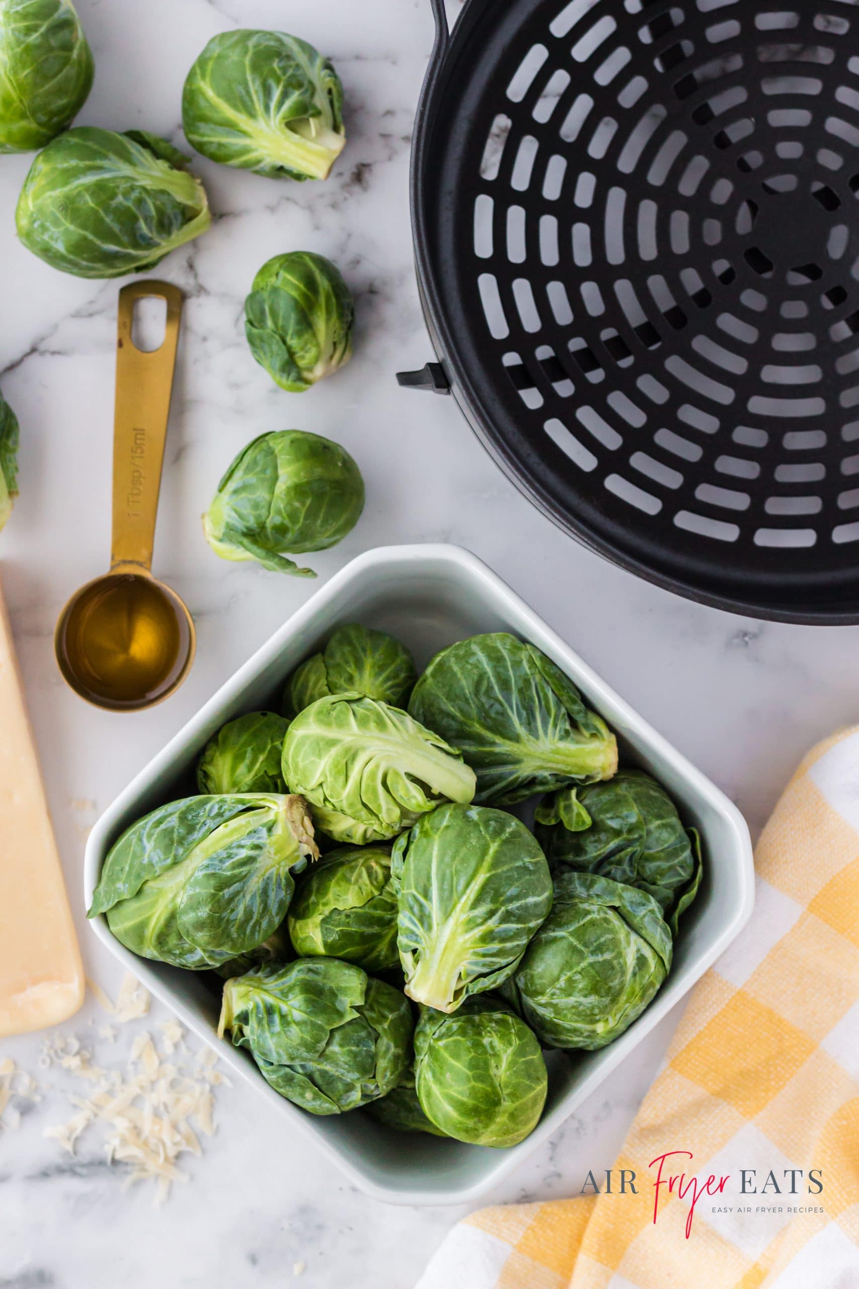 A square bowl of fresh raw brussel sprouts next to a gold tablespoon of oil and the air fryer basket for a Ninja Foodi