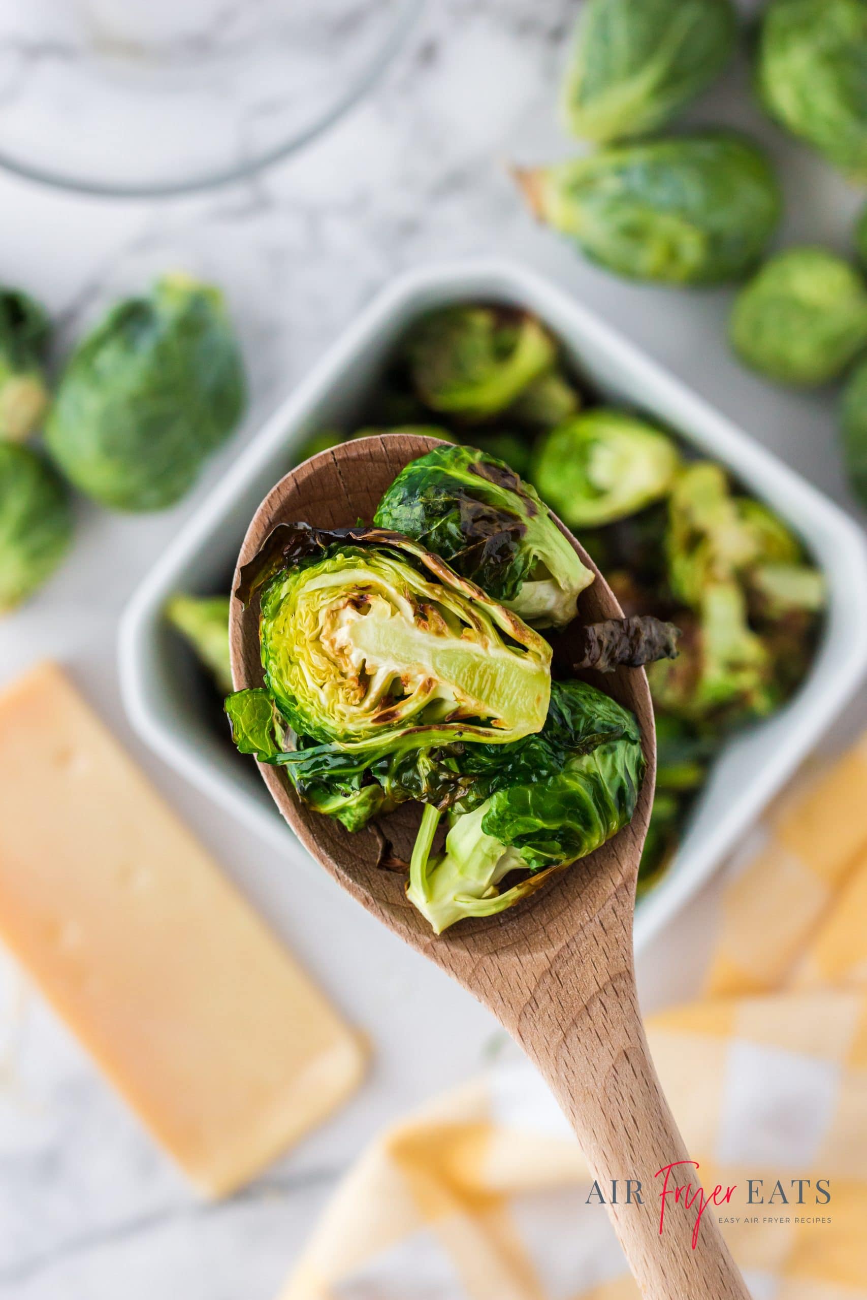 a wooden spoon holding a few halved, roasted brussel sprouts above a square serving bowl of them.