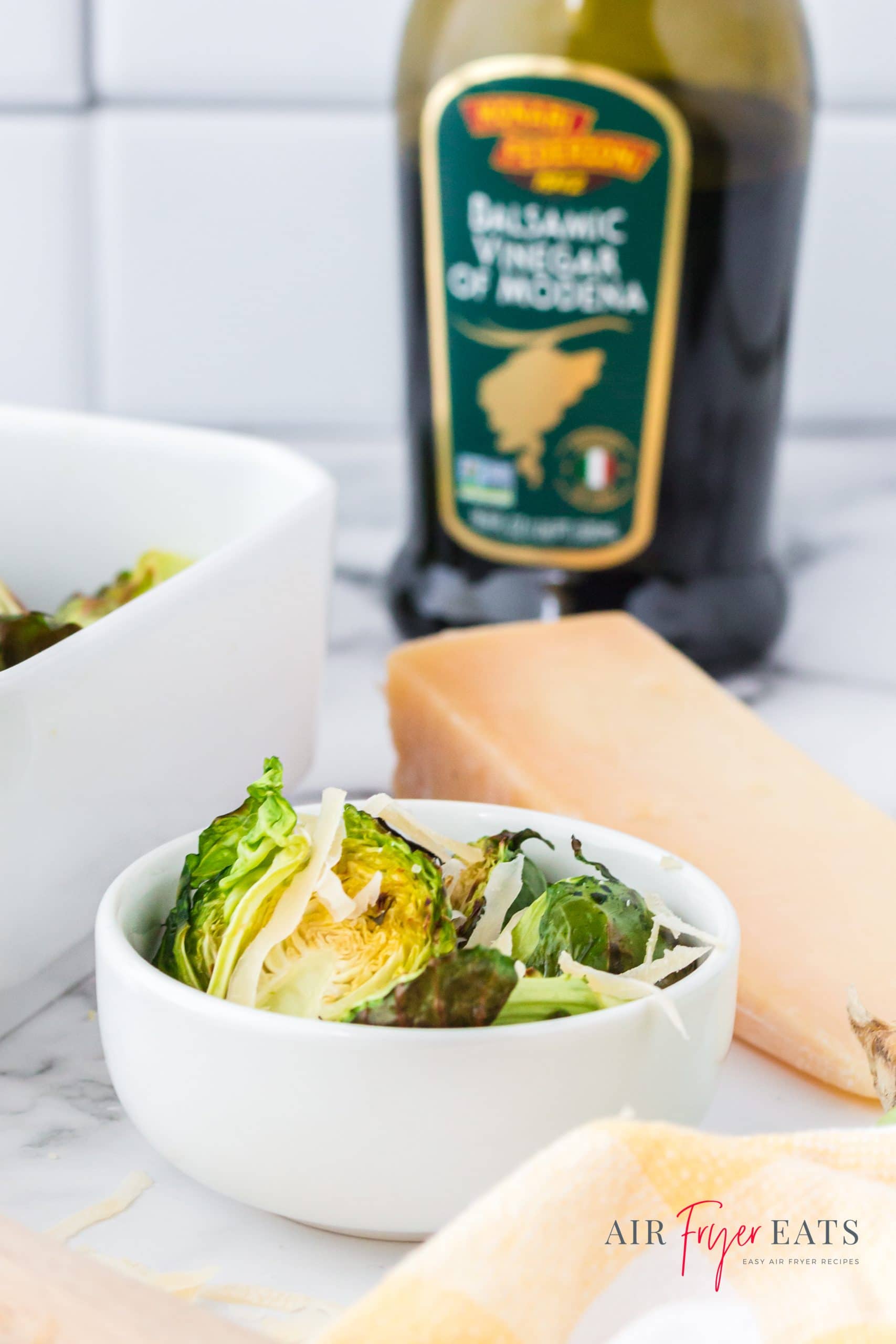 a bowl of air fried brussel sprouts topped with balsamic and parmesan. A bottle of balsamic vinegar is in the background