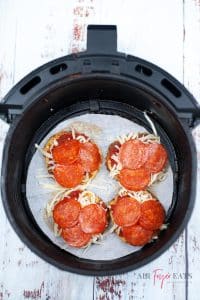 A black air fryer basket containing 4 air fried cauliflower pizzas with cheese and pepperoni on top of them