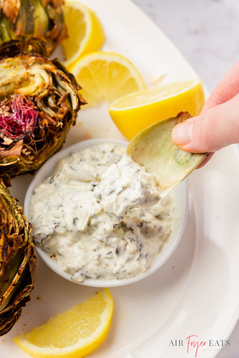 a hand dipping an artichoke leaf into a dipping sauce