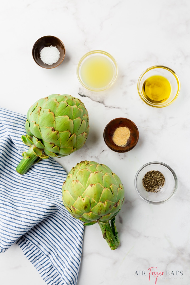 Two artichokes, oil, and seasonings on a marble counter, viewed from above