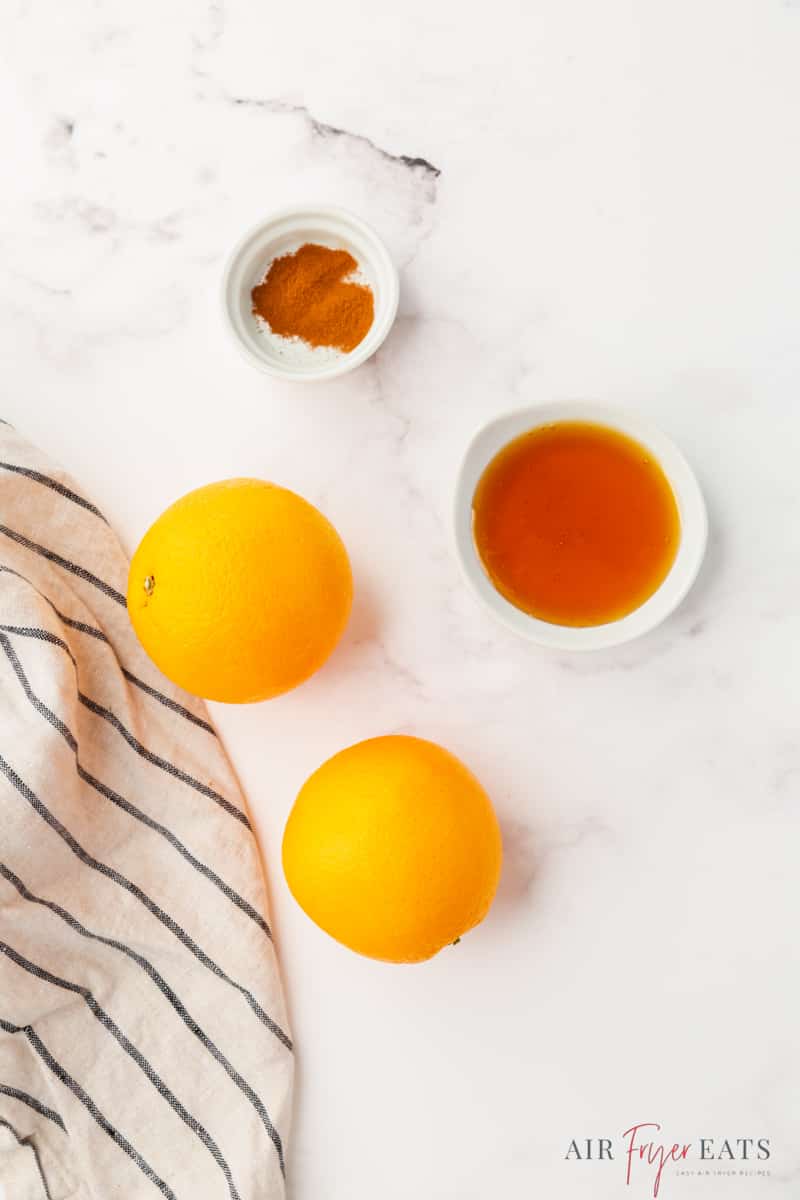 two oranges, a small bowl of cinnamon, and a small bowl of maple syrup on a marble counter, viewed from above