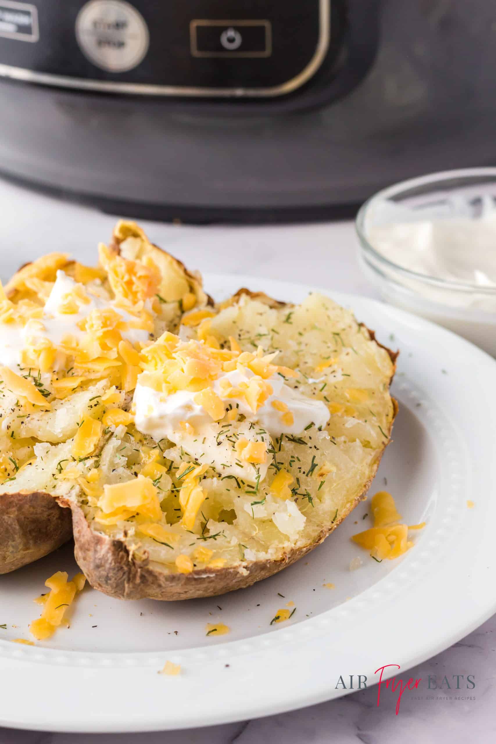 a split baked potato on a white plate, sitting in front of a ninja foodi. The potato is topped with sour cream, cheese, and seasonings