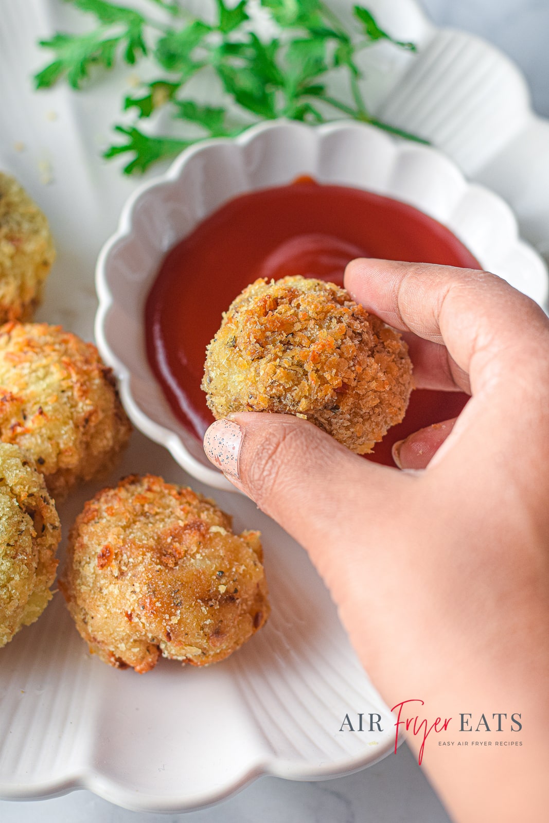 A white plate containing air fried cheesy potato balls, and a hand lifting a ball up to take a bite