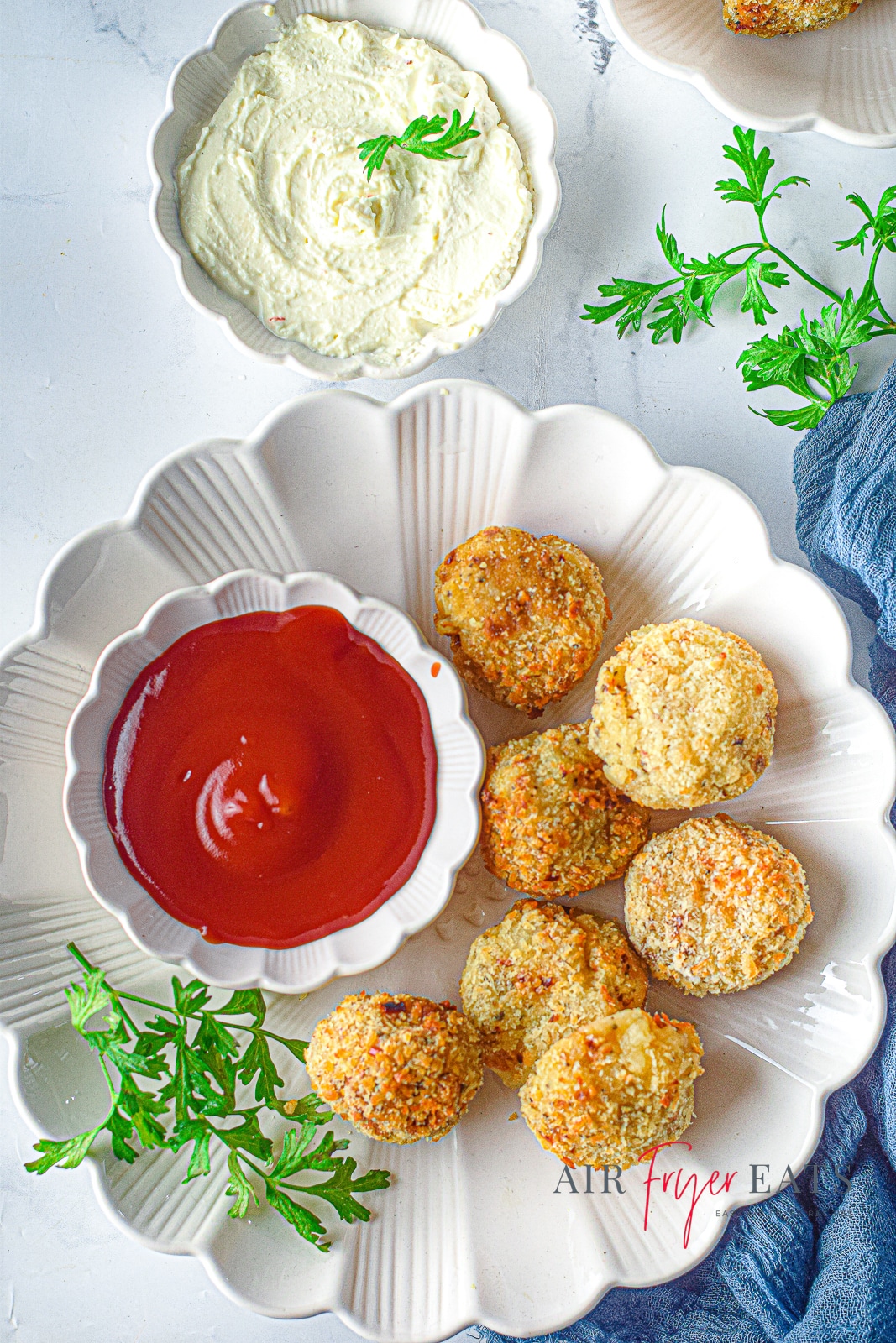 Birds eye view of a white plate containing air fried cheesy potato balls. Also on the plate is a white ramekin containing red sauce. Around the plate is a bowl of creamy white mashed potato