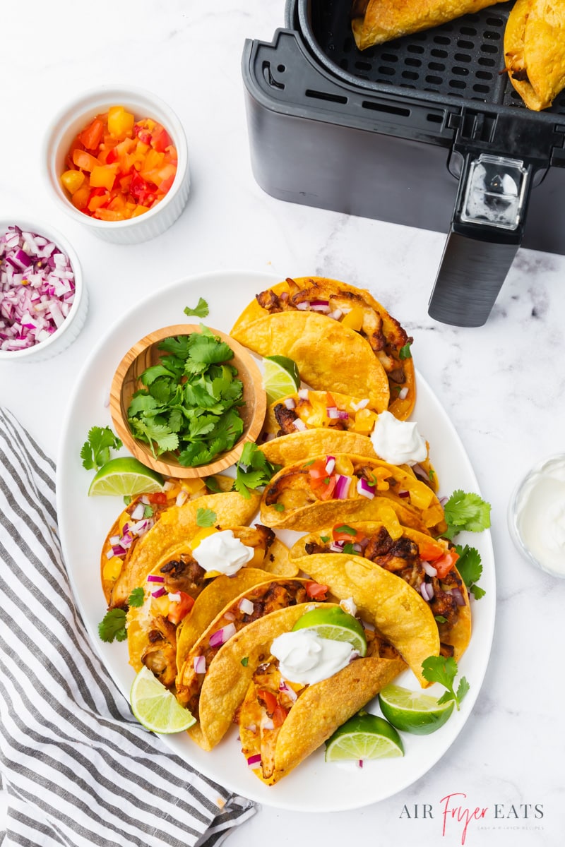 a platter of tacos topped with pico, sour cream, lime wedges, next to an open air fryer, with a bowl of pico de gallo and chopped onions.