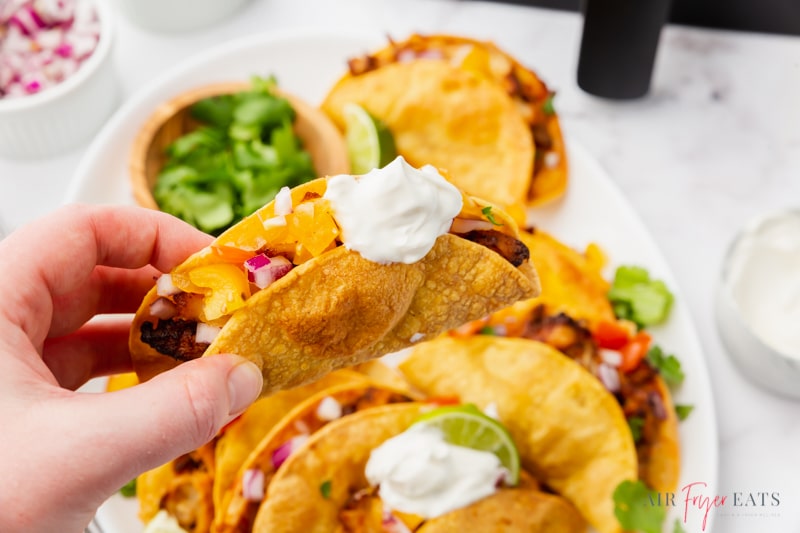 a hand holding an air fryer taco above a platter of tacos. The taco has been dipped into sour cream