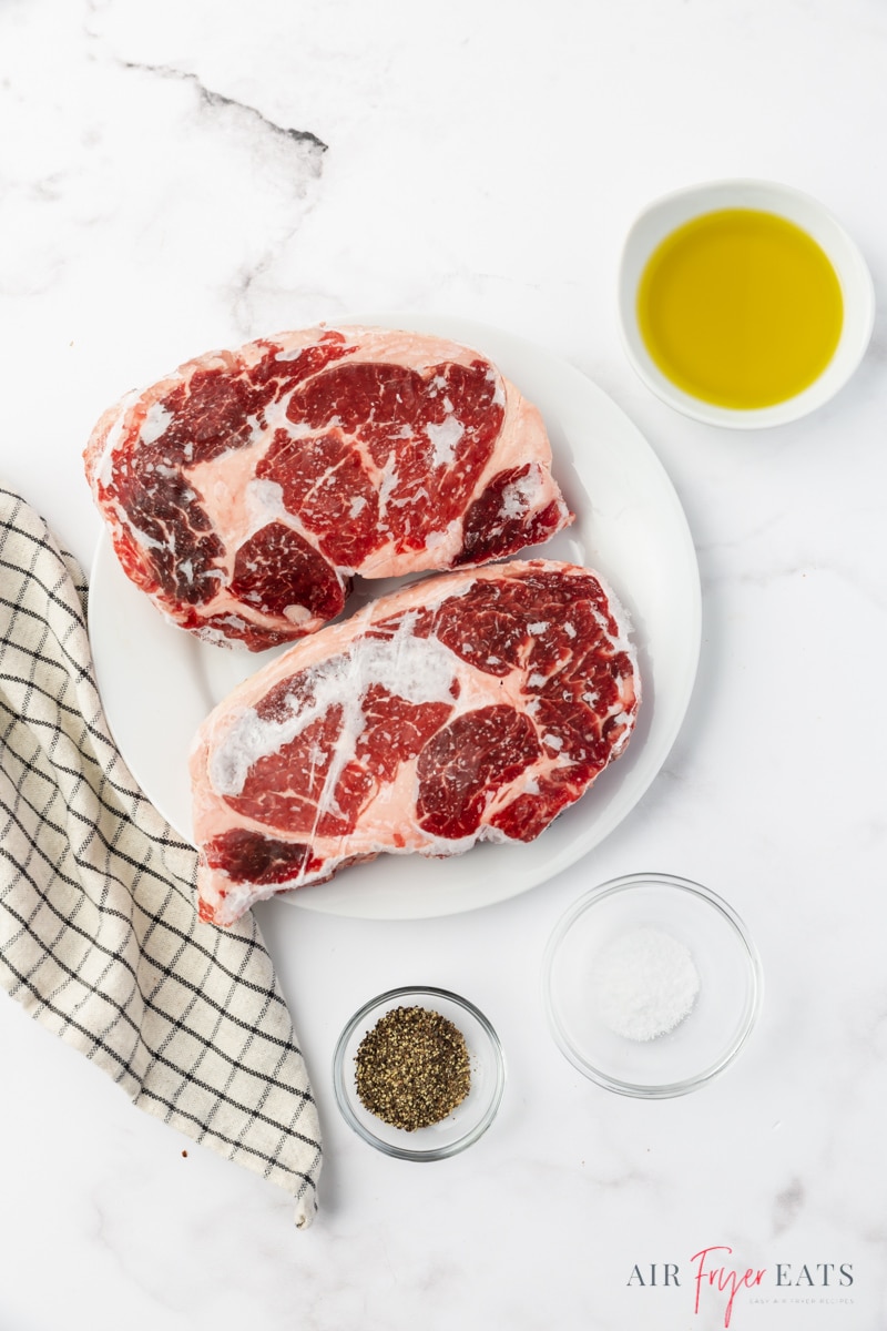 two frozen steaks on a white plate, with salt, pepper, and olive oil.