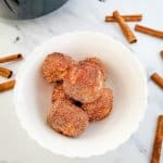 Vertical photo of 6 golden brown donut holes on a white plate. Around the plate are cinnamon sticks and in the background there is a black air fryer with silver handle