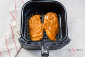 A black air fryer basket containing two chicken breasts coated in mustard mixture