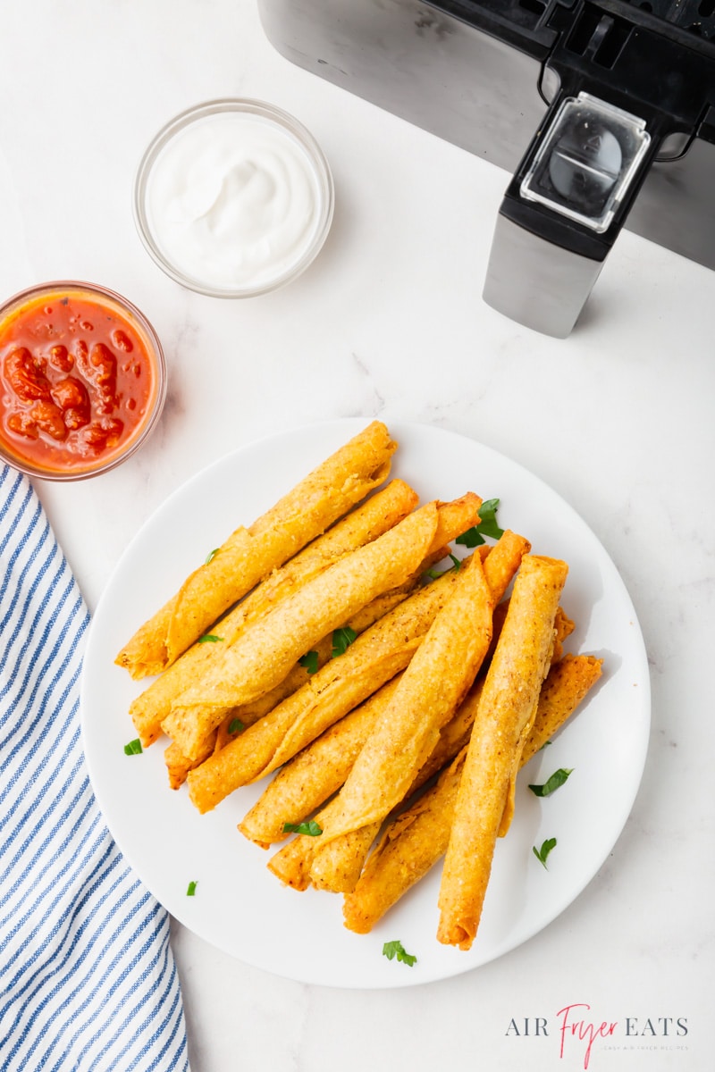 A plate of taquitos next to an air fryer with a small bowl of salsa and another of sour cream, viewed from overhead