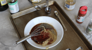 A white bowl with mustard, soy sauce and spices being mixed with a whisk. Around the bowl are the ingredients required for the maple dijon dressing. Dijon mustard, maple syrup, soy sauce, minced garlic, Italian seasoning, smoked paprika & oil