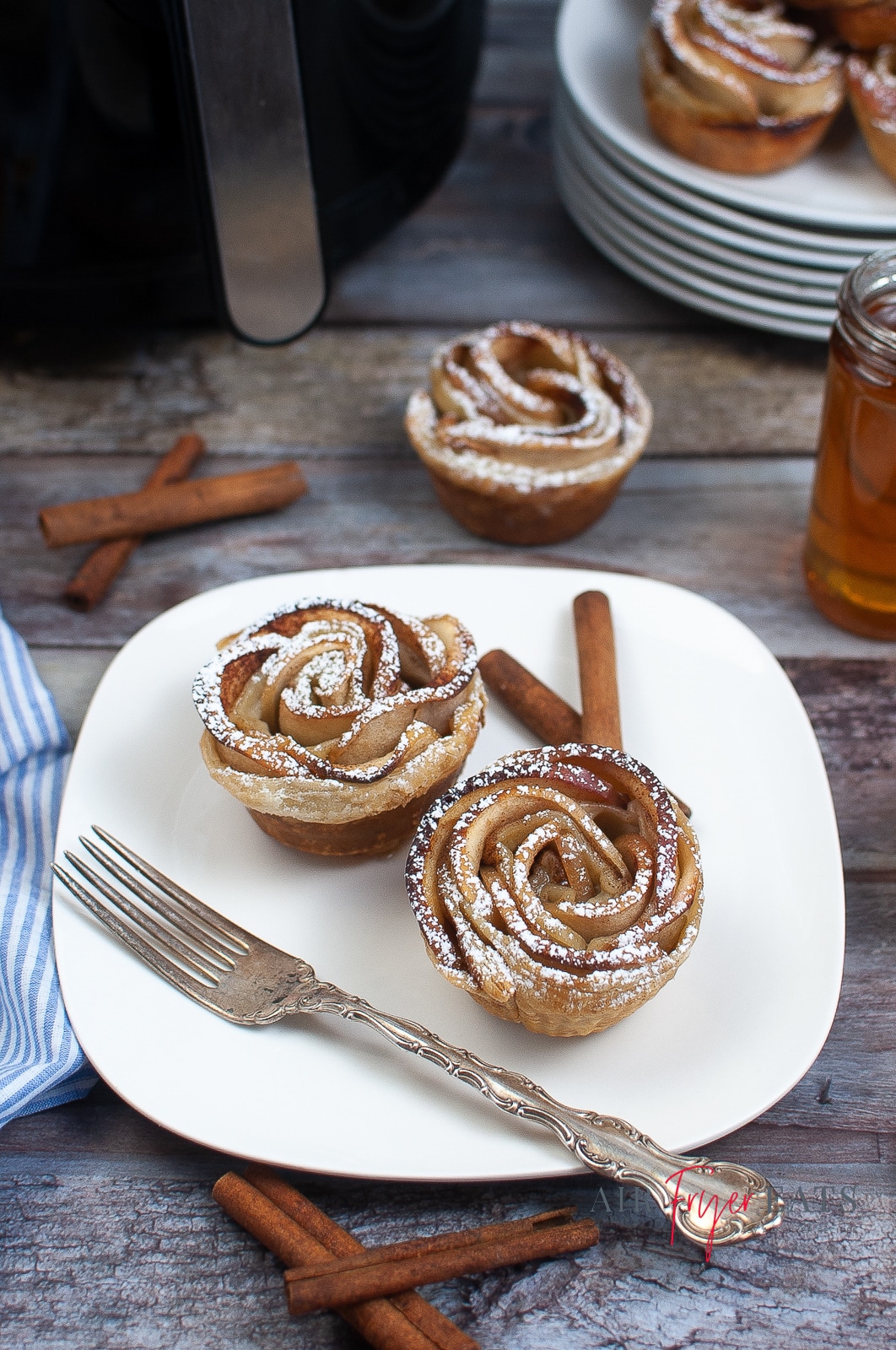 A white plate containing two air fried apple roses and a silver fork. There are other apple roses and ingredients scattered around the outside of the plate