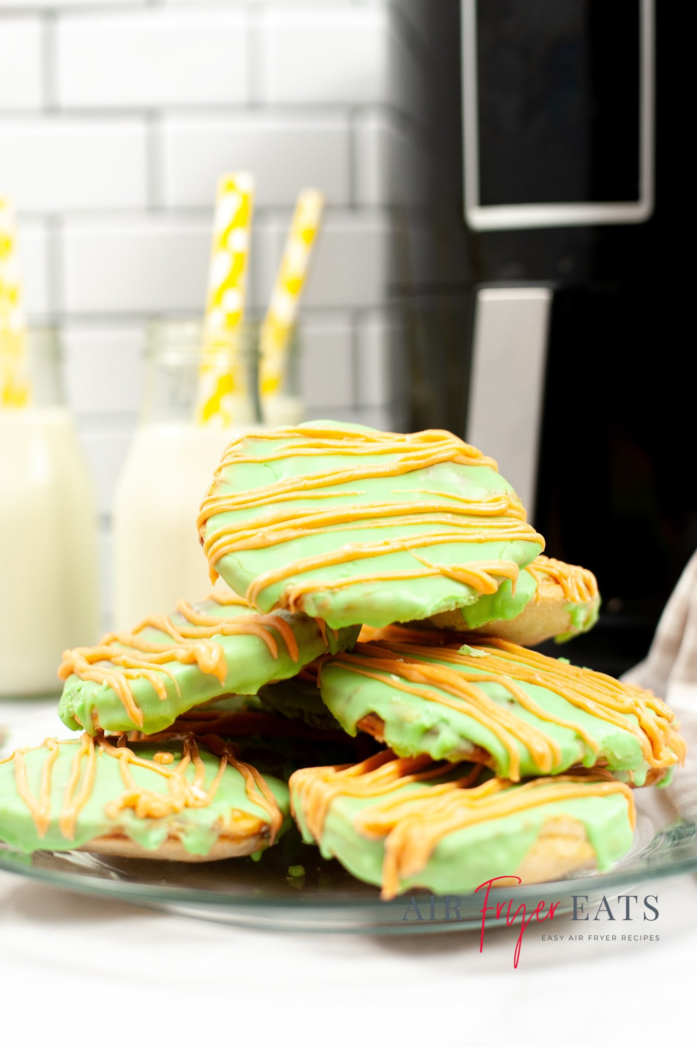 Vertical photo of a pile of green poptarts on a plate. In the background is a black air fryer and bottles of milk