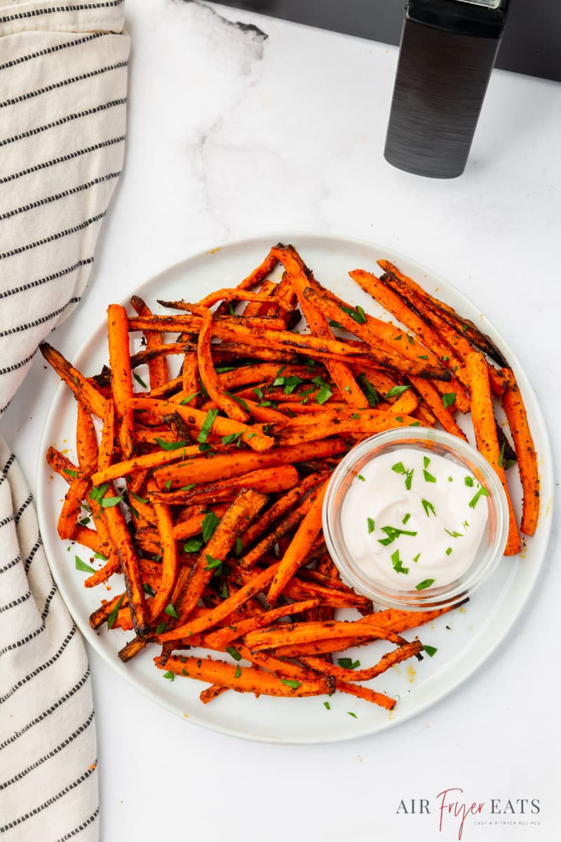 a round plate of air fryer carrot fries with creamy dipping sauce on the side, in front of an air fryer basket