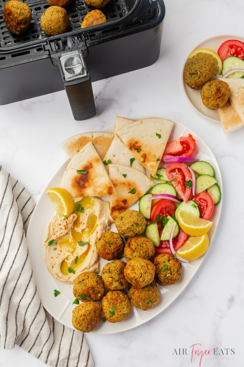 an oval platter of air fryer falafel, cucumber and tomato salad, and pita, next to an air fryer basket.
