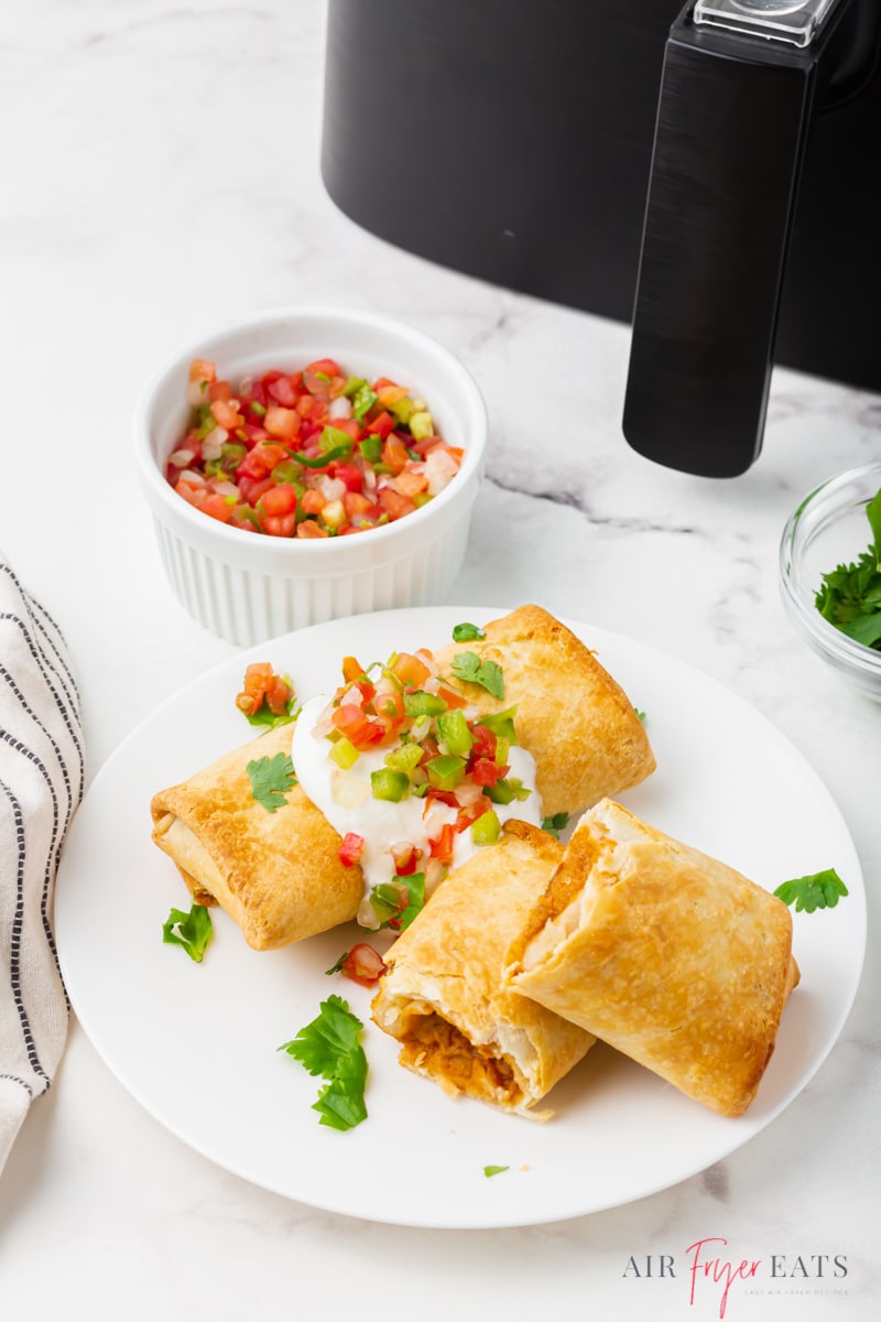 two crispy chimichangas topped with sour cream, salsa, and cilantro. in the background is a cup of salsa and an air fryer.