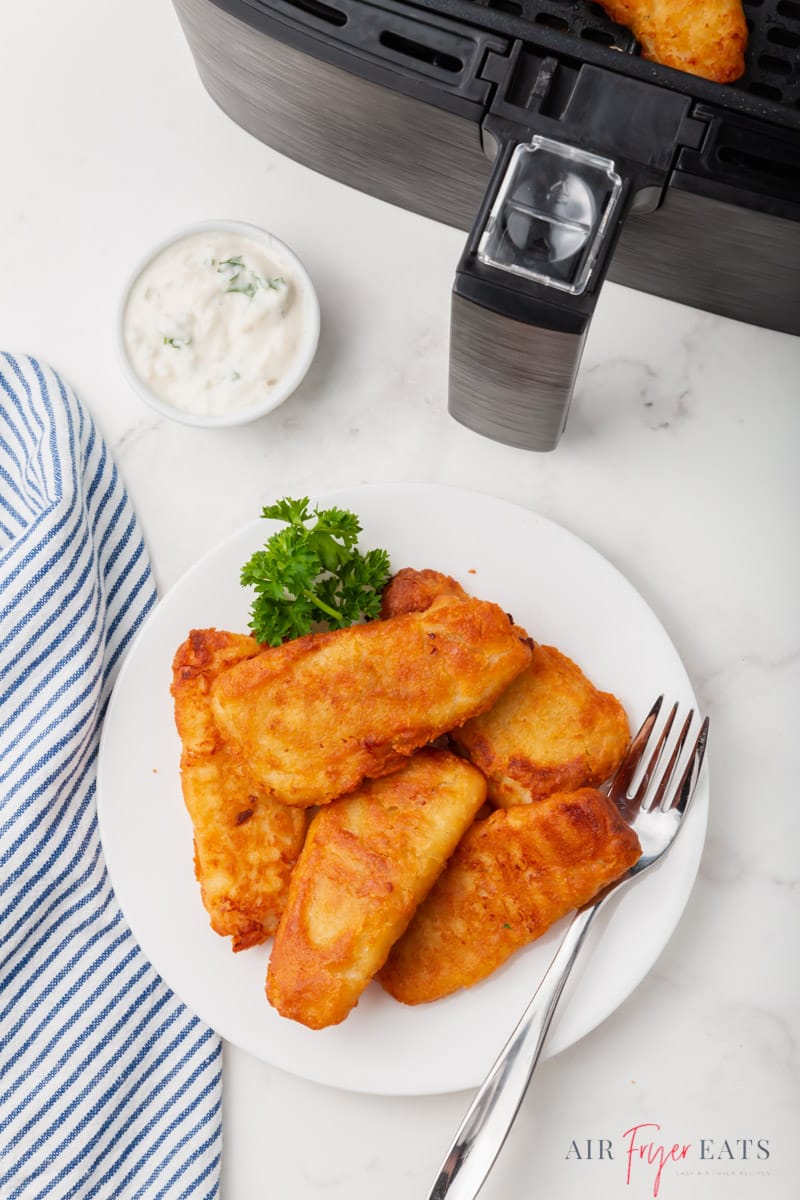 a plate of cooked frozen fish fillets in front of an air fryer basket on a marble counter. 
