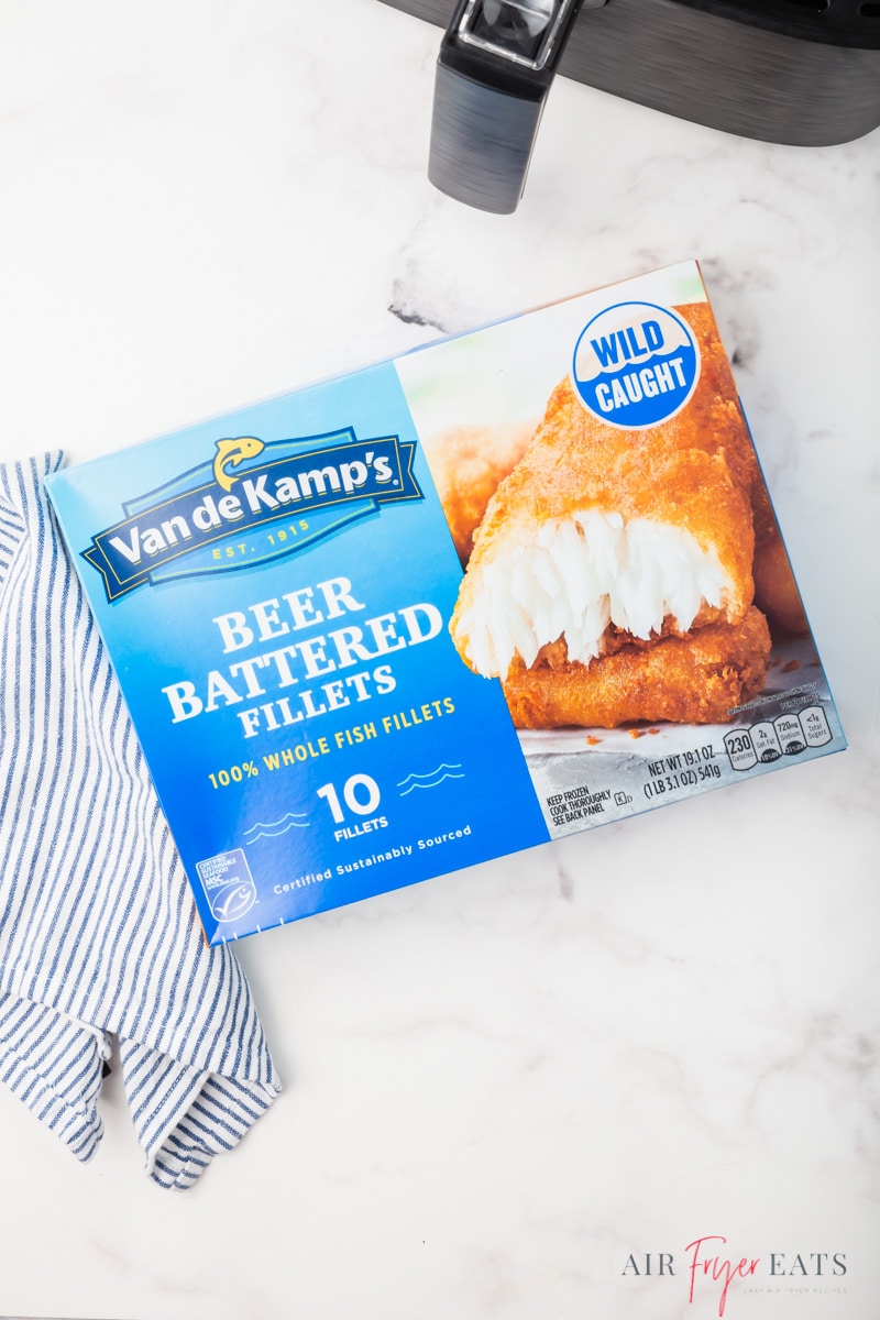 a box of van de kamps beer battered fish fillets on a counter next to a striped towel. the handle of an air fryer basket is in the top right corner of the shot. 
