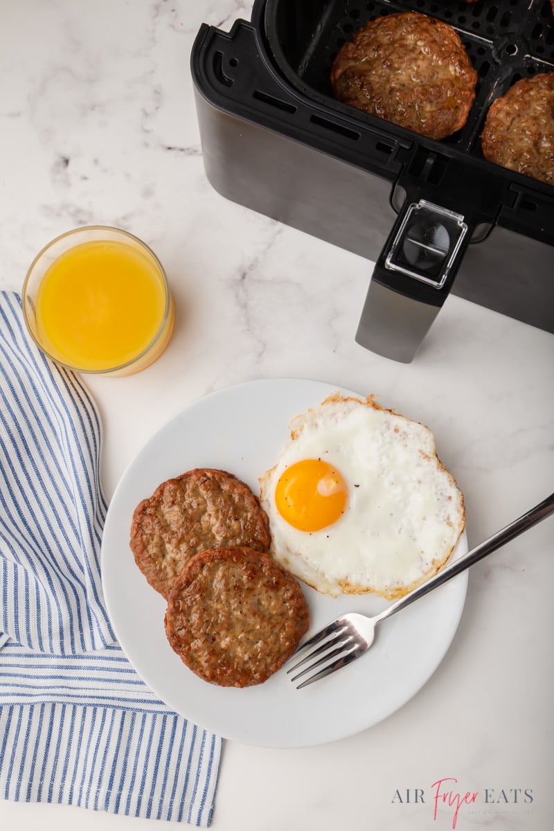 a white plate with one sunny side egg and two large sausage patties. There's a fork on the plate, a glass of orange juice next to it. To the right is a cosori air fryer basket with sausage patties in it.