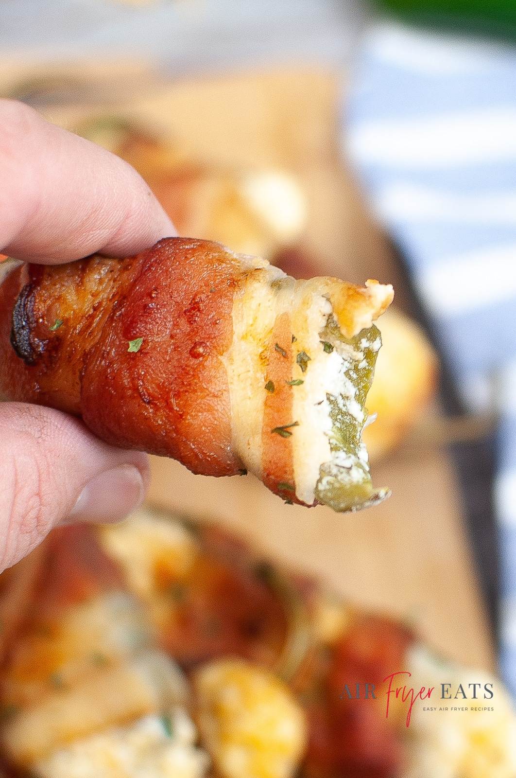 vertical photo of somebody holding a bacon wrapped jalapeno popper, with some other poppers on a board underneath