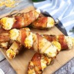 vertical photo of 7 bacon wrapped jalapeno poppers on a board, which is on a wooden table with a striped cloth, a jalapeno pepper and some grated cheese