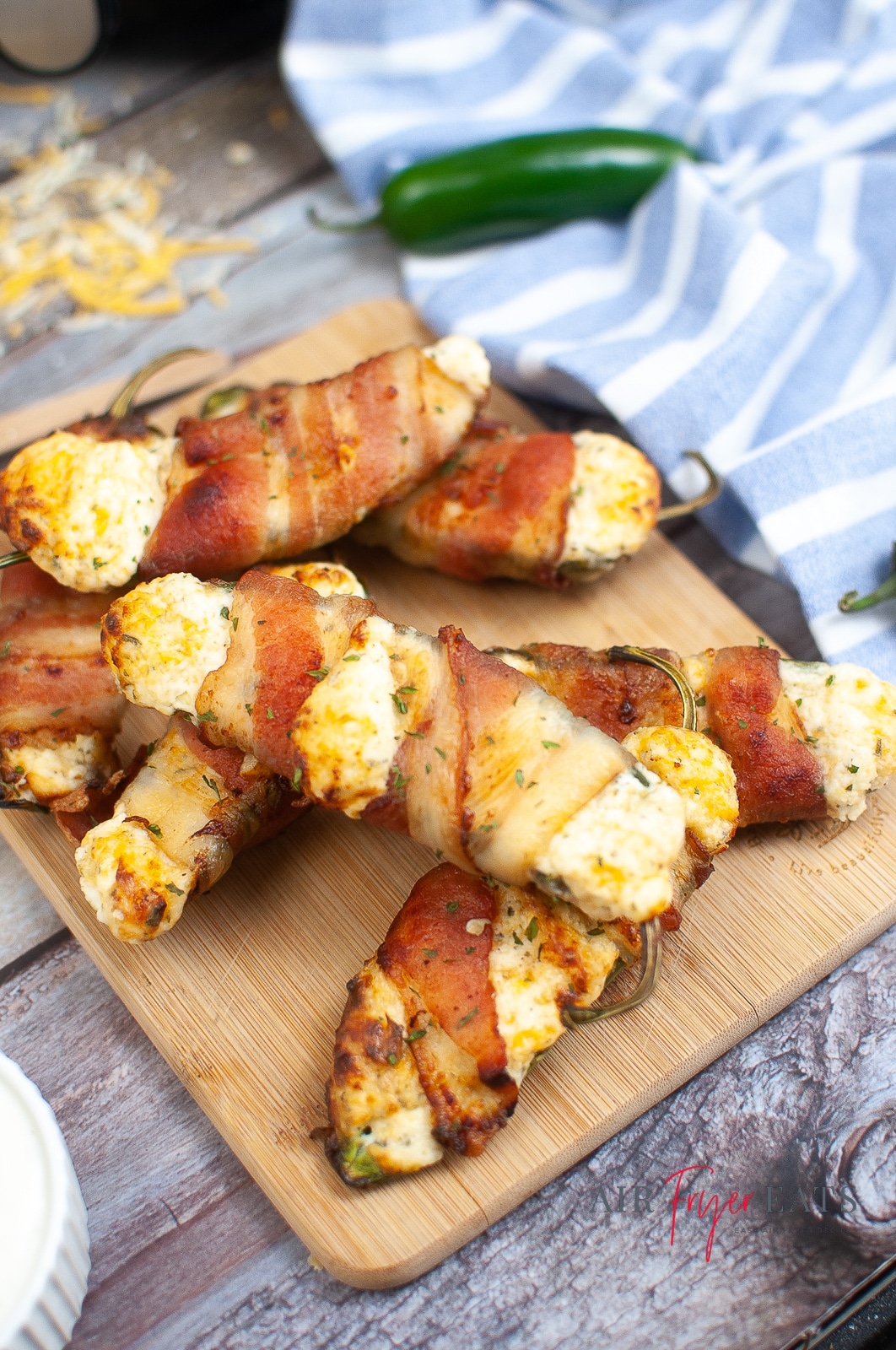 vertical photo of 7 bacon wrapped jalapeno poppers on a board, which is on a wooden table with a striped cloth, a jalapeno pepper and some grated cheese are also on the table