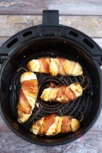 vertical photo of 4 air fryer bacon wrapped jalapeno poppers in a basket type air fryer on a wooden table