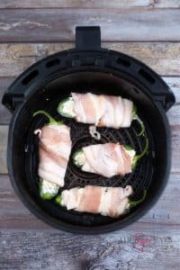 vertical photo of air fryer bacon wrapped jalapeno poppers in air fryer basket, ready to cook