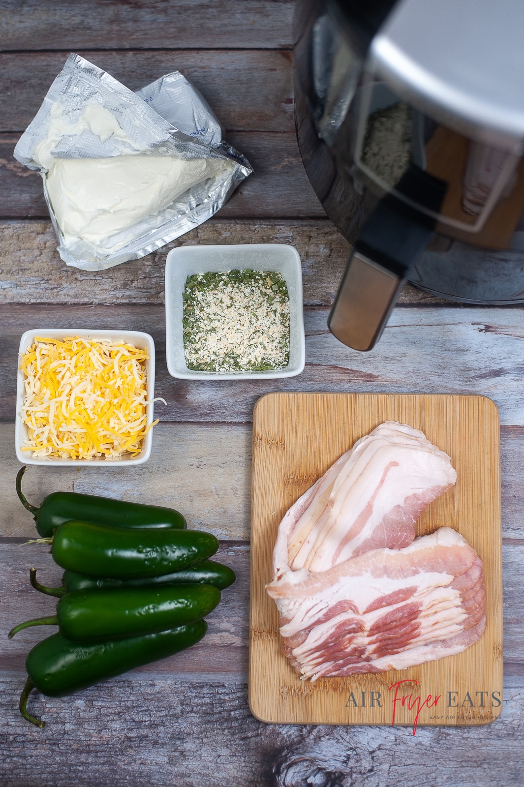 vertical photoof air fryer bacon wrapped jalapeno poppers ingredients. Cream cheese, shredded cheese, jalapeno peppers bacon and ranch seasoning on a wooden table with an air fryer in the top corner