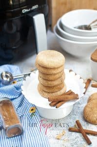 A stack of golden brown coloured snickerdoodles on a white plate with some cinnamon sticks to the side. In the background is various dishes and utensils that have been used. There is also a black air fryer with silver handle