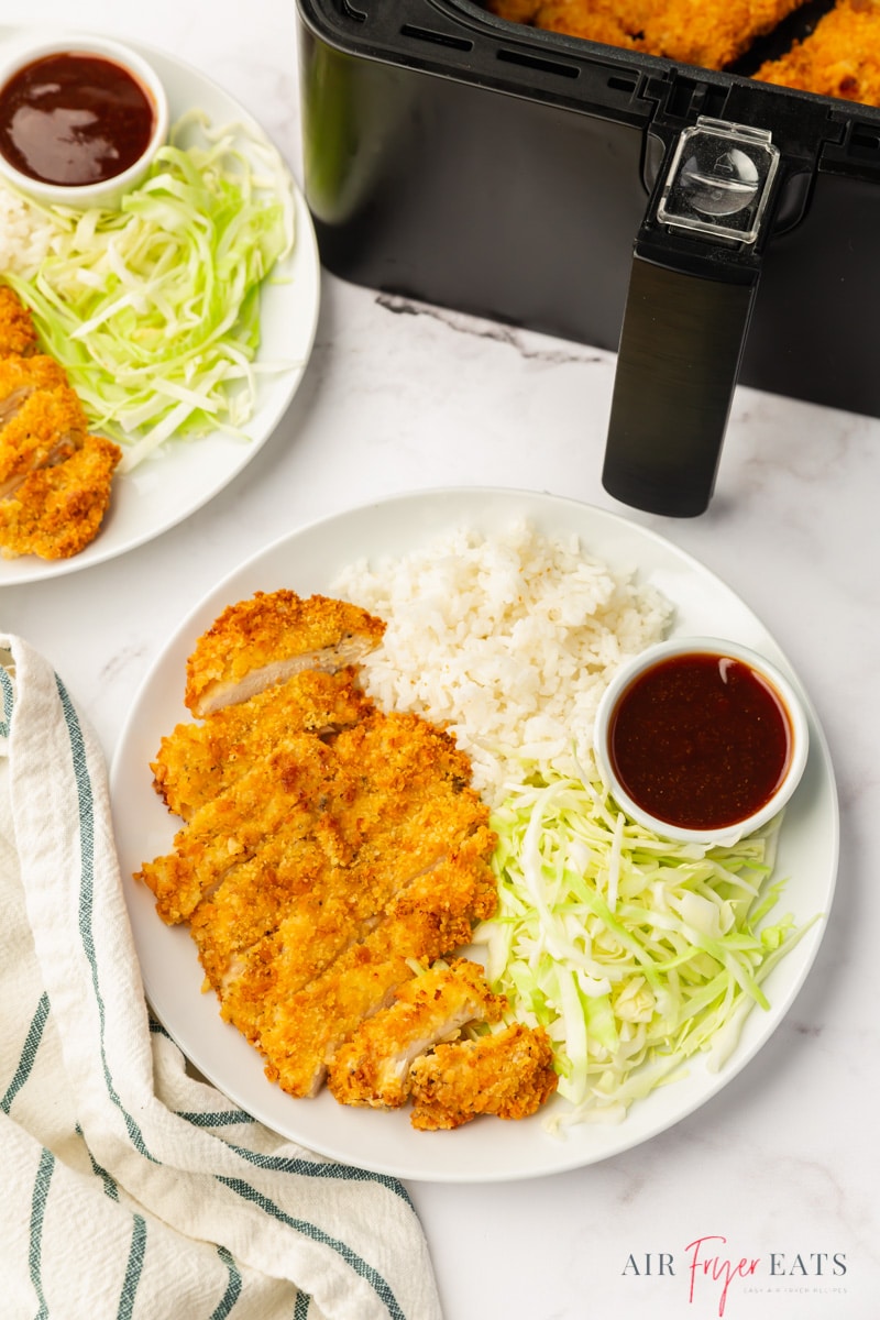 dinner plates next to an air fryer basket. Each plate has a sliced crispy chicken katsu cutlet with white rice, shredded