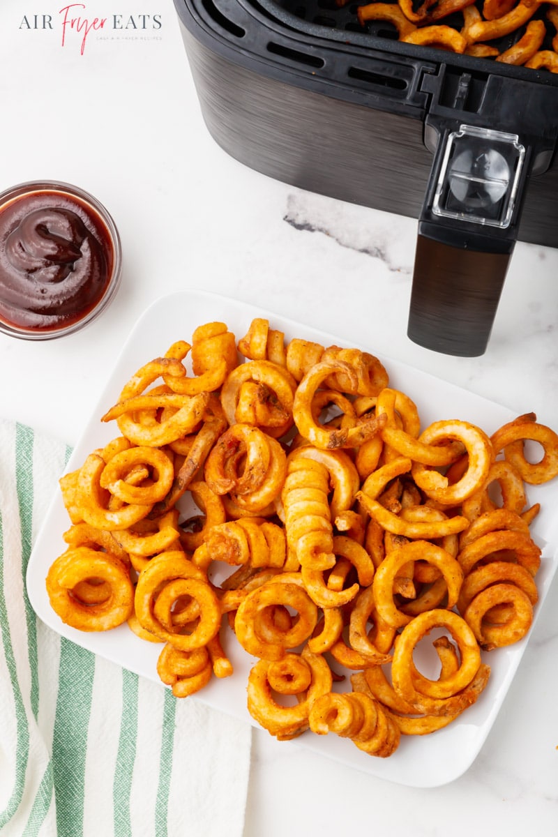 a plate of curly fries in front of an air fryer. a side of bbq sauce is on the side.